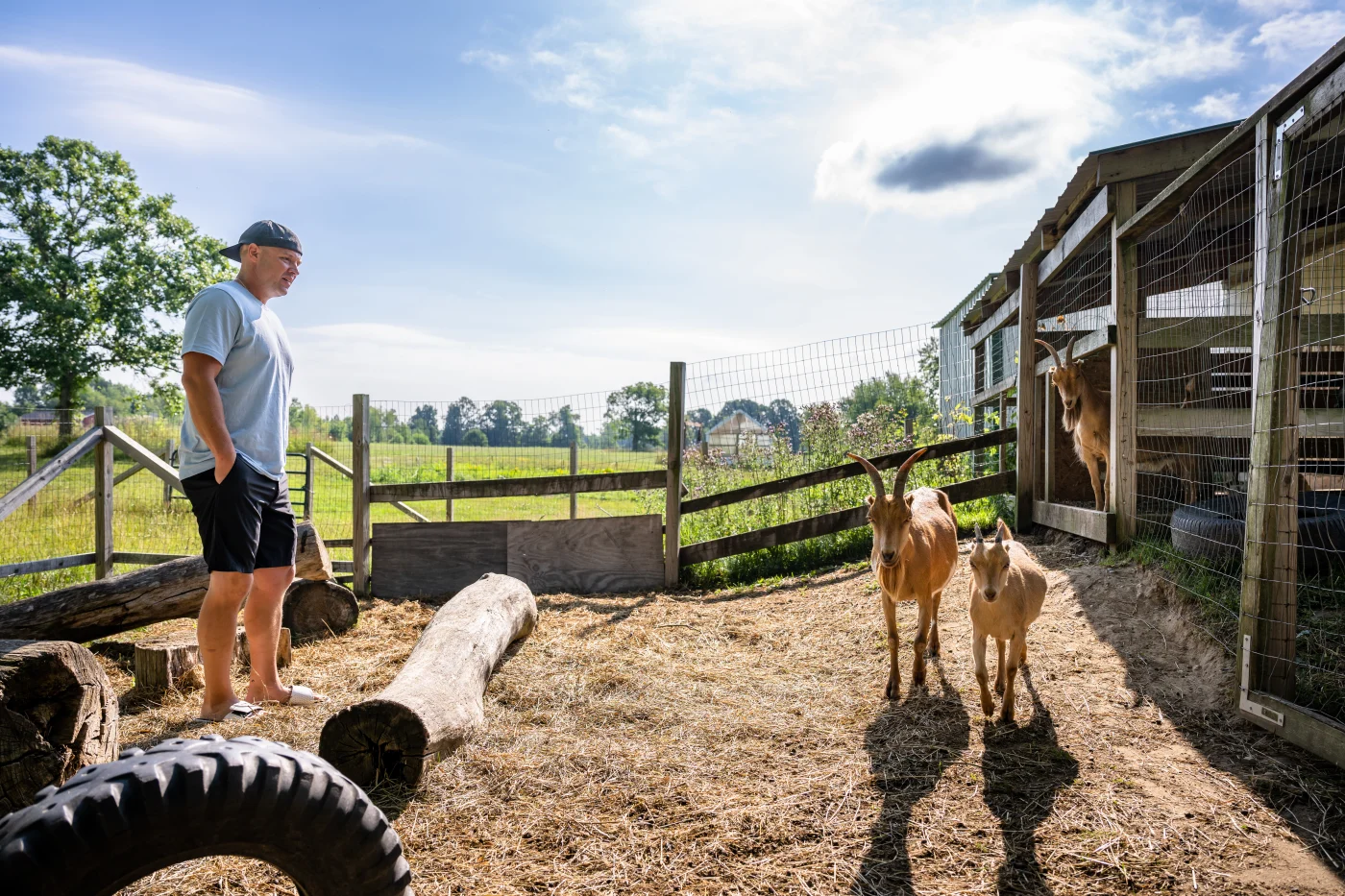 A man standing in a goat pen on a family farm wearing dark blue shorts, a light blue t-shirt, and a backward hat, looking at two goats in front of him.